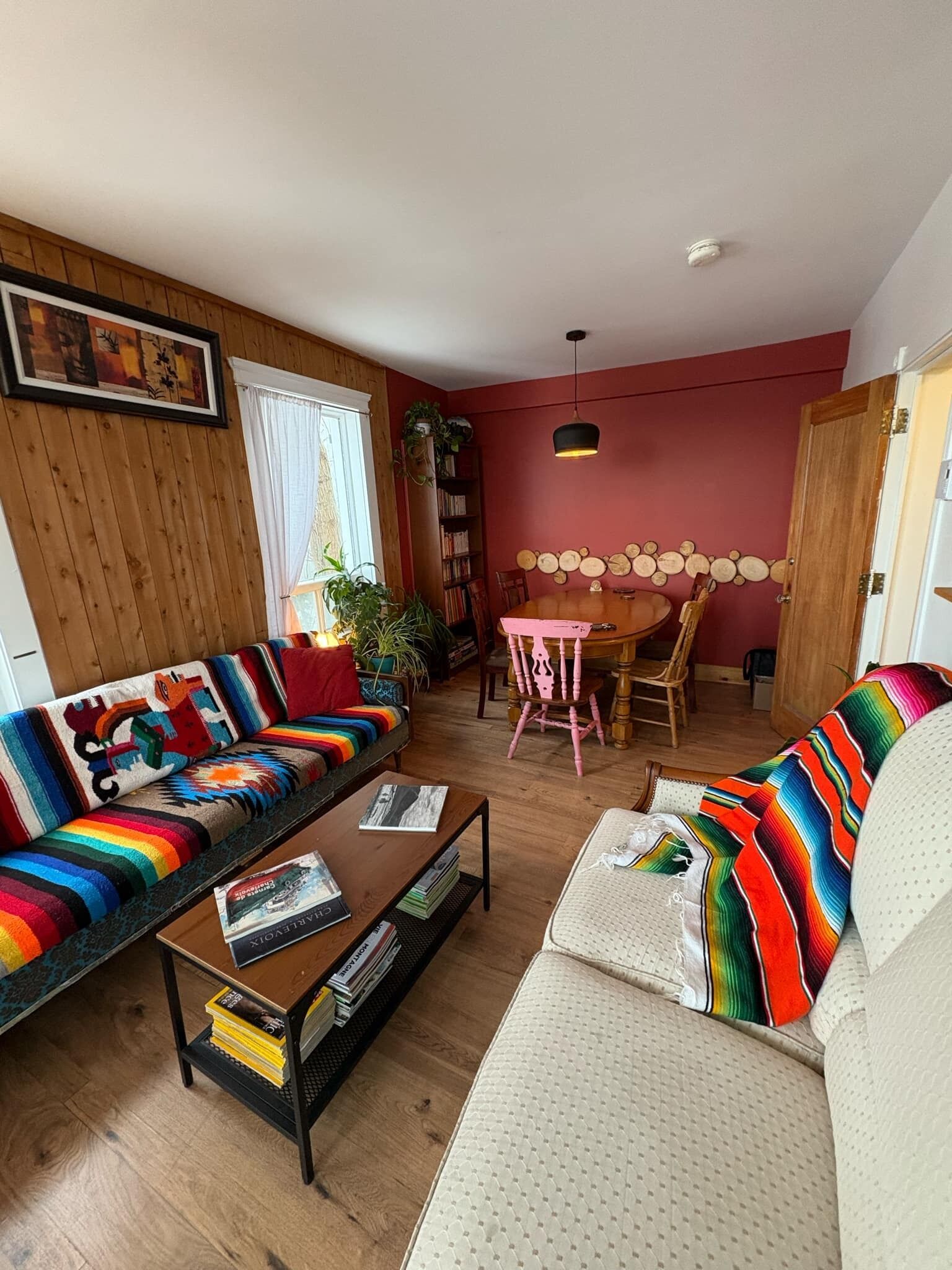 A cozy living room with a wooden floor, a colorful sofa, a white sofa, a wooden coffee table, and a dining table with chairs.