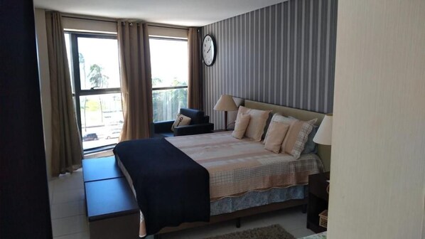 Un dormitorio con cama, mesitas de noche, una silla, un ventanal con cortinas y un reloj en la pared.