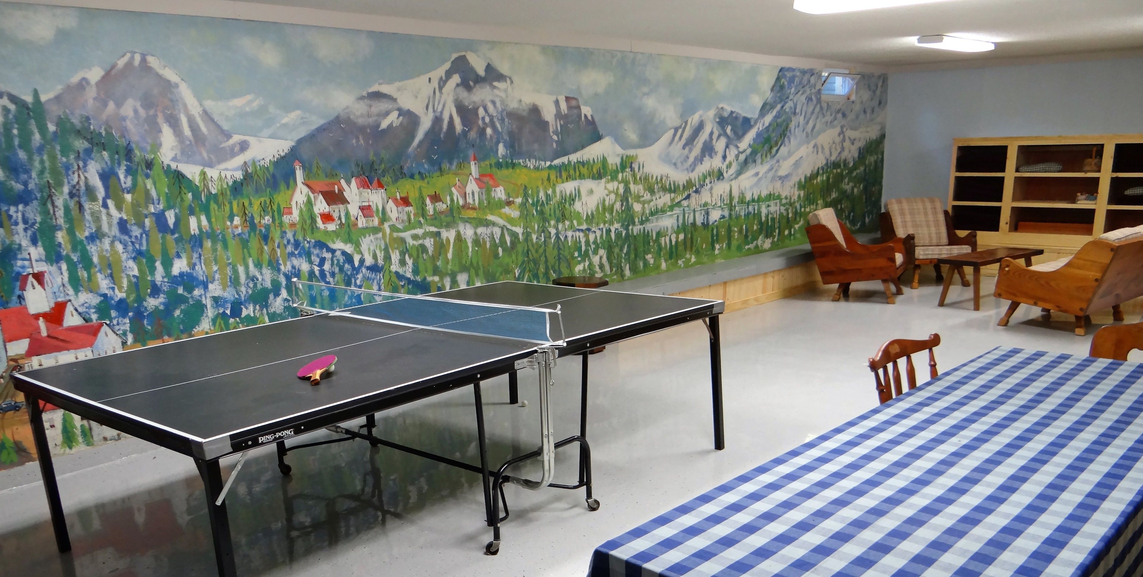 A ping pong table in a room with a mountain landscape mural, a checkered tablecloth, and wooden chairs.
