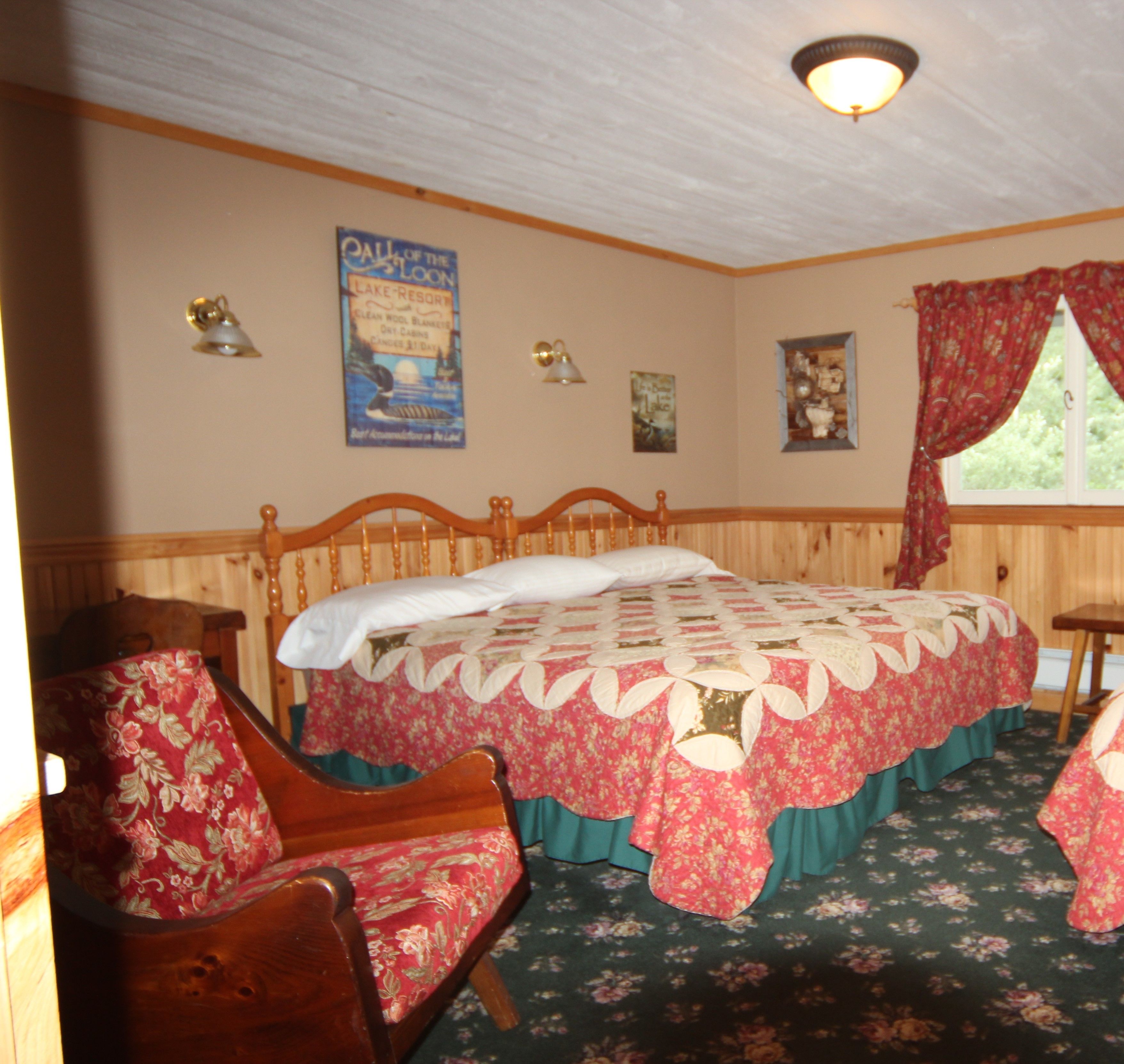 A bedroom with a wooden bed, floral-patterned chairs, a quilt, a poster on the wall, and a window with curtains.