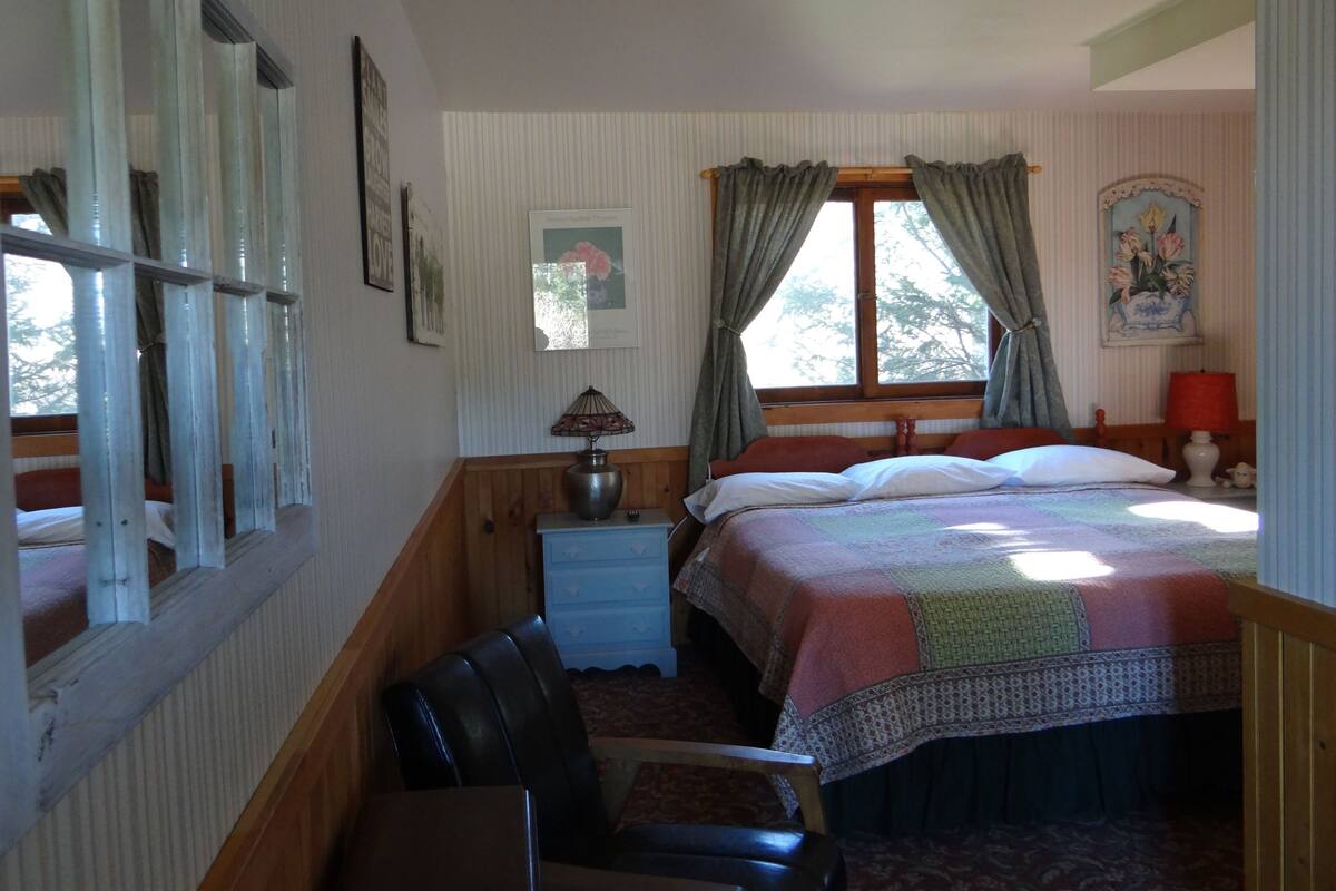 A bedroom with a bed, a nightstand, a chair, a window with curtains, and a wall with framed pictures.