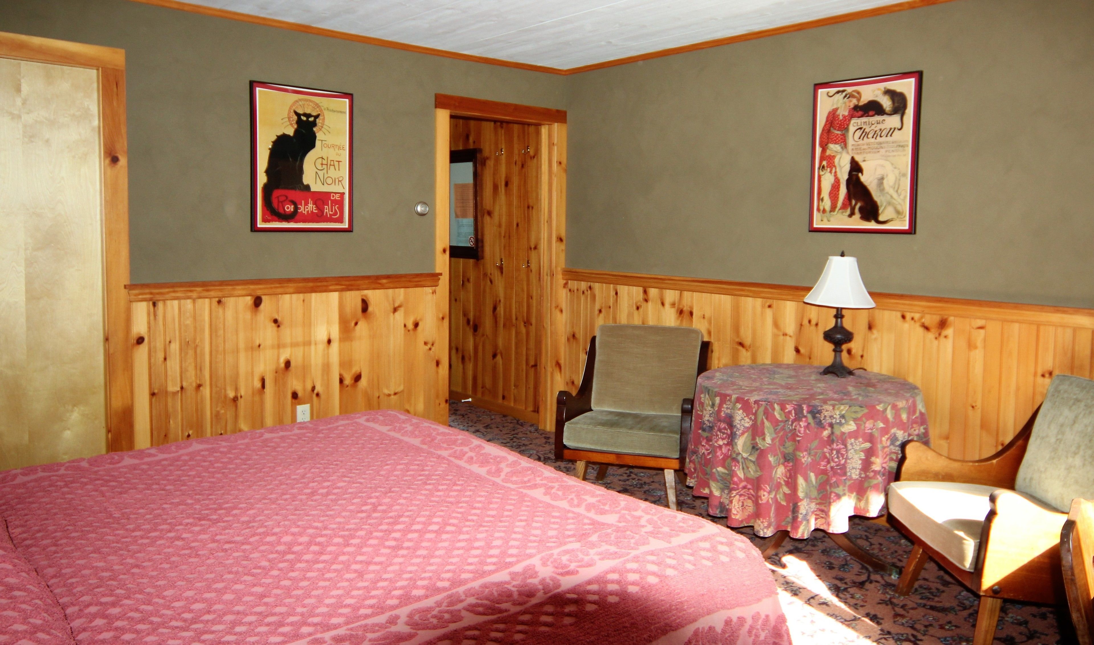 A room with a bed, two chairs, a table with a lamp, and two posters on the wall.
