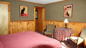 A room with a bed, two chairs, a table with a lamp, and two posters on the wall.