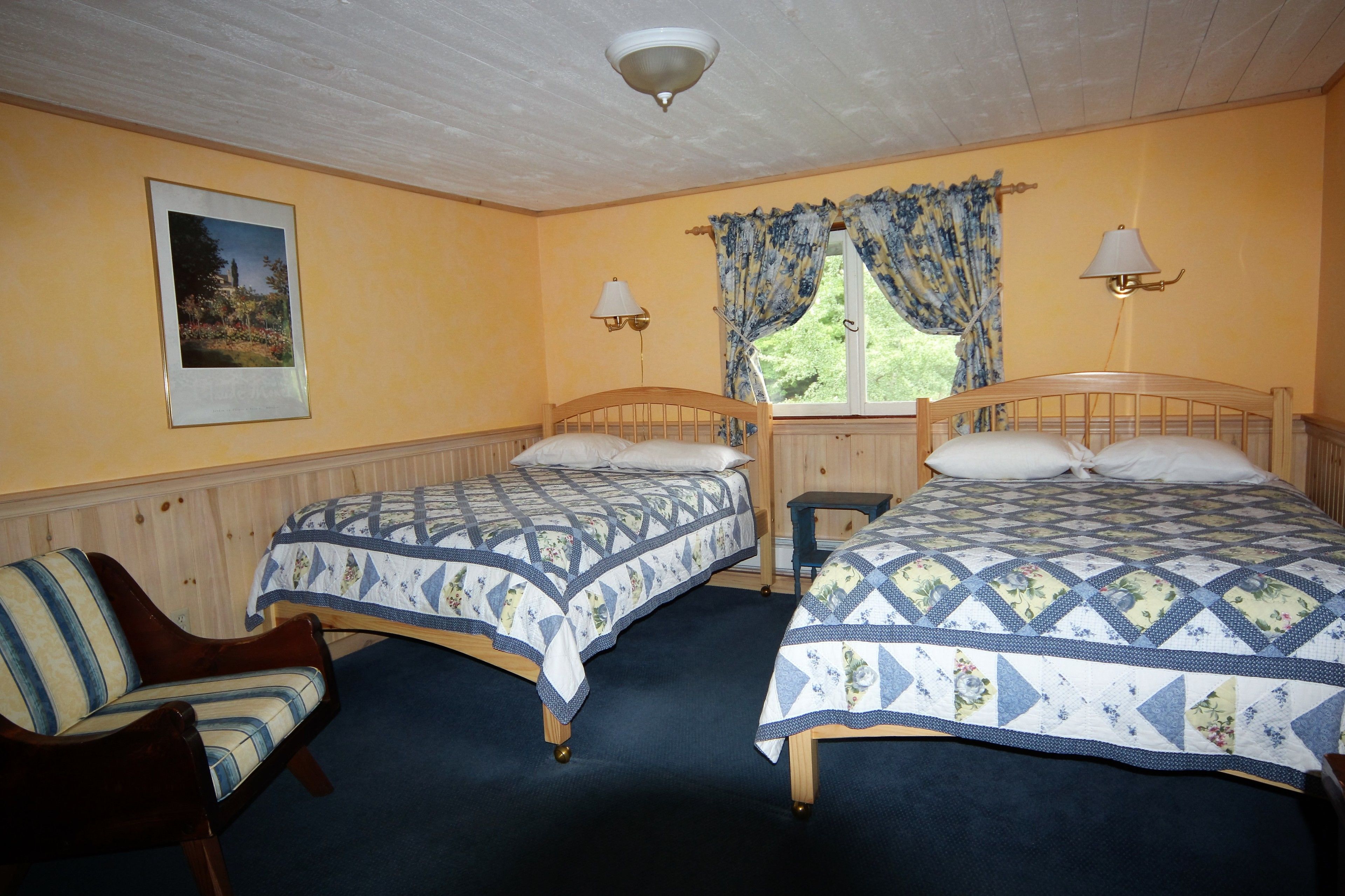A room with two wooden beds, a chair, a window with curtains, a wall-mounted lamp, and a framed picture.