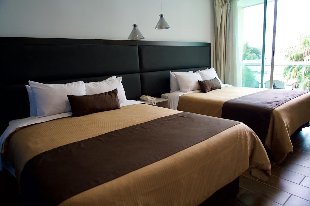A hotel room with two beds, a dark headboard, bedside tables, and a view of the outdoors.