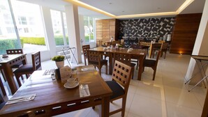 A modern dining area with wooden tables and chairs, a kitchen in the background, and large windows providing natural light. - Hotel Posada Señorial (San Andrés Cholula)