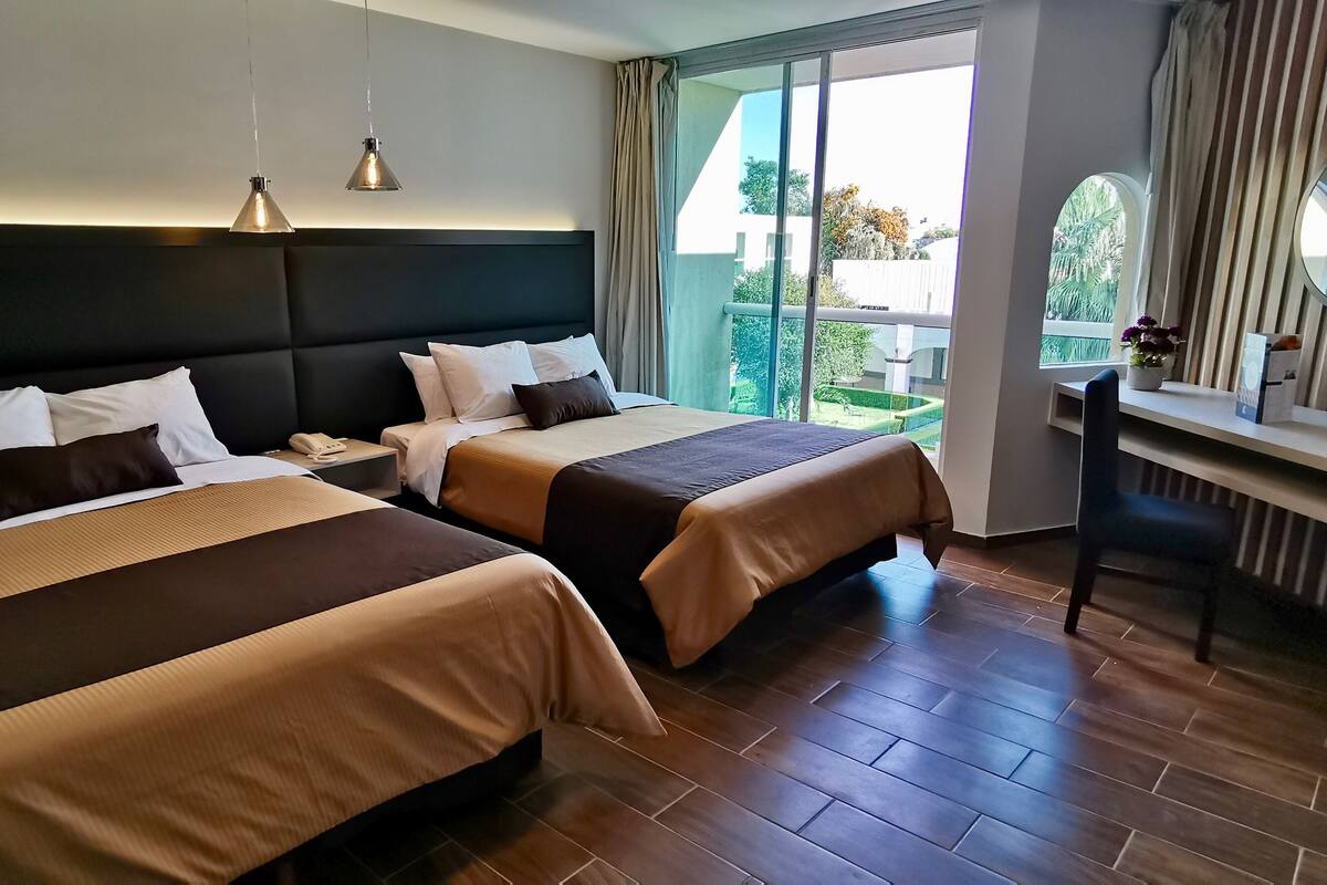 A hotel room with two beds, a desk, a mirror, and a large window.