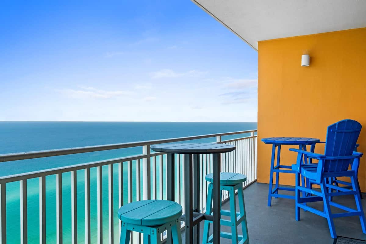 A balcony with turquoise chairs and a table, overlooking a clear blue sea.