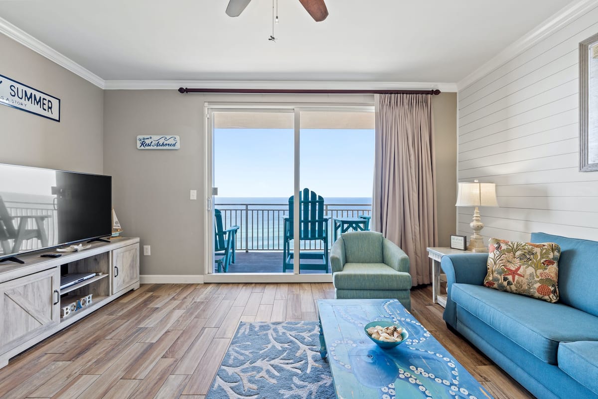 A living room with a TV, a sofa, a coffee table, and a view of the ocean through a sliding glass door.