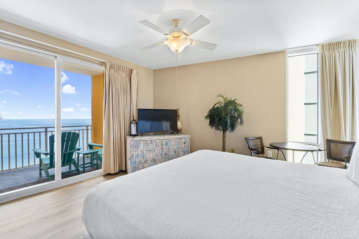 A hotel room with a large bed, a ceiling fan, a TV on a cabinet, a small table, two chairs, a potted plant, and a balcony with a view of the ocean.