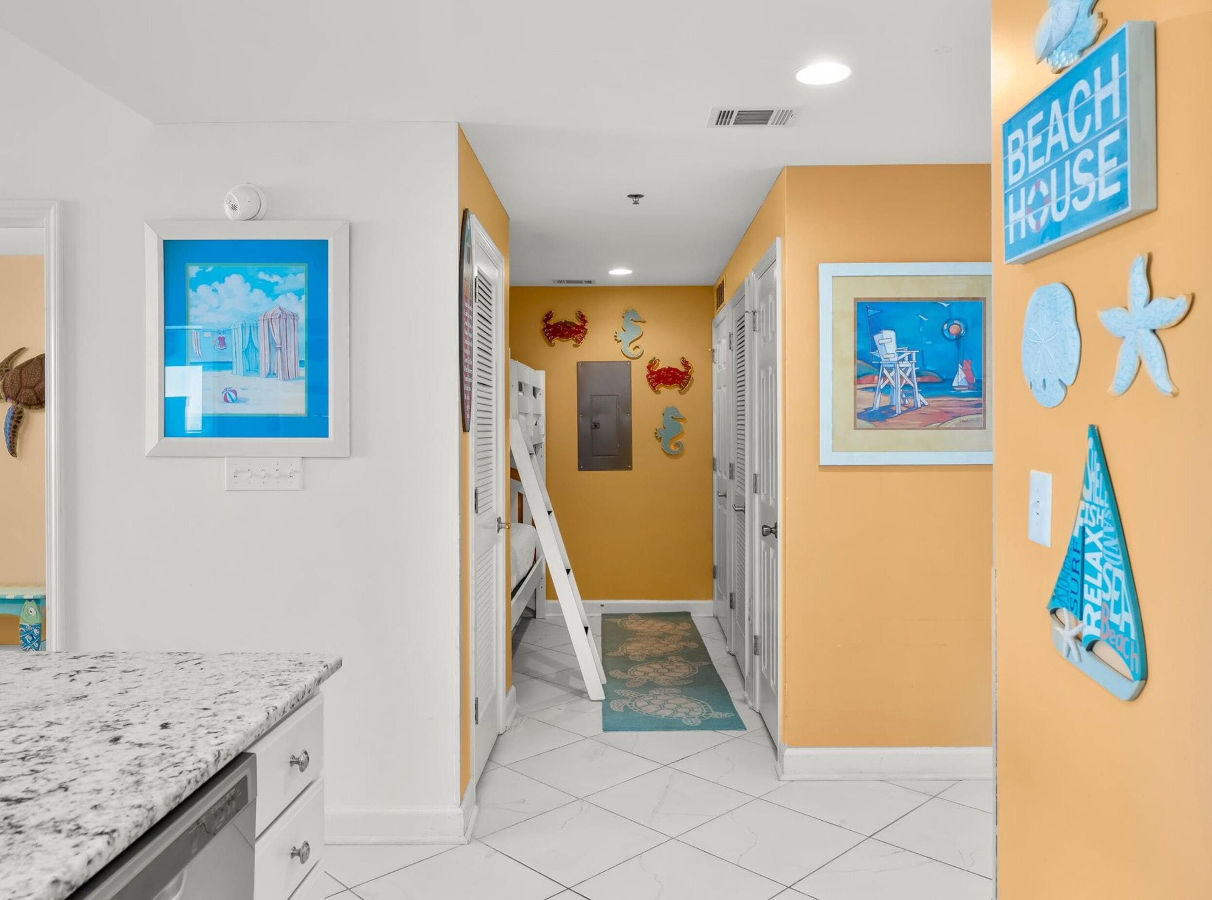 A narrow hallway with a kitchen area on the left, featuring a granite countertop and white cabinets. The walls are decorated with beach-themed artwork and wall hangings.