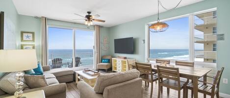 A spacious living room with a large window overlooking the ocean, a ceiling fan, and a dining area with wooden furniture.