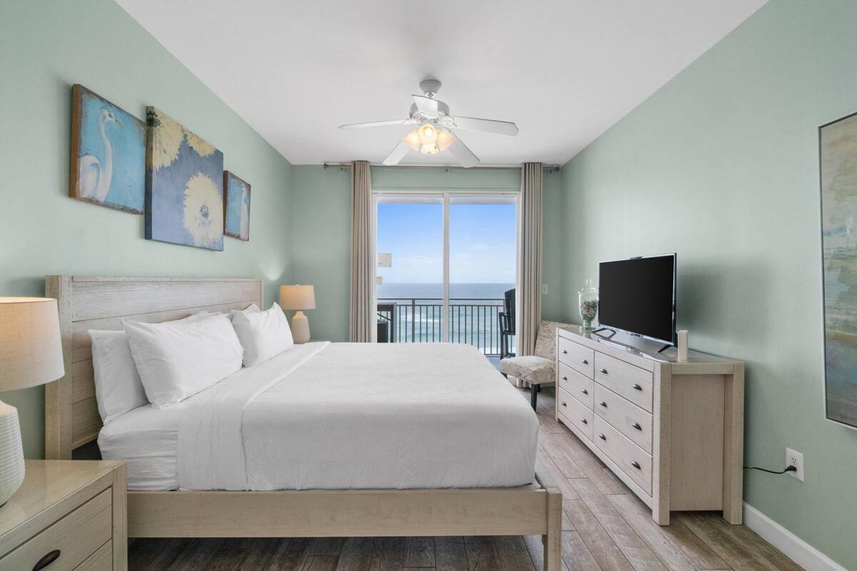 A bedroom with a bed, a TV, a ceiling fan, and a view of the ocean through a large window.