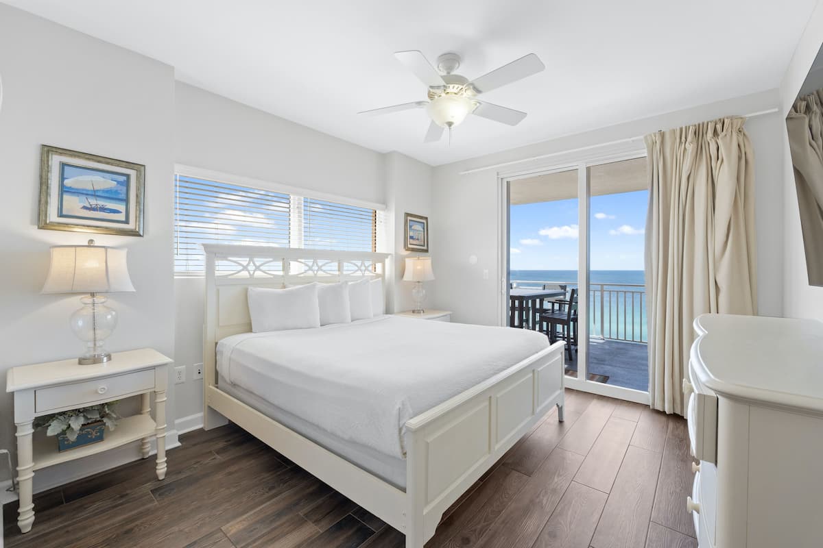 A bedroom with a bed, two bedside tables, a ceiling fan, and a view of the ocean through large sliding doors.