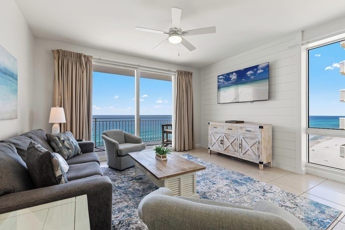 A living room with a sofa, armchair, coffee table, and a view of the ocean through large windows.