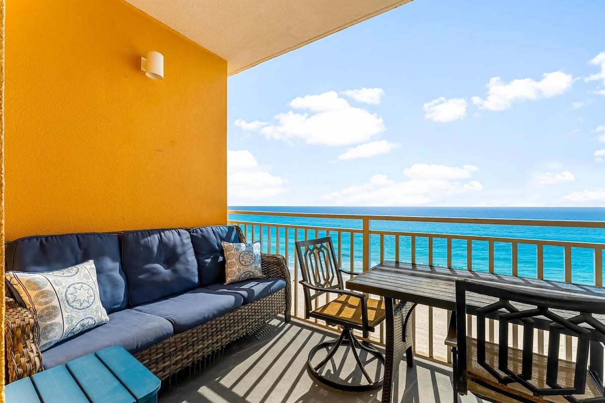 A balcony with a wicker sofa, a wooden table, and a chair, overlooking a turquoise sea.