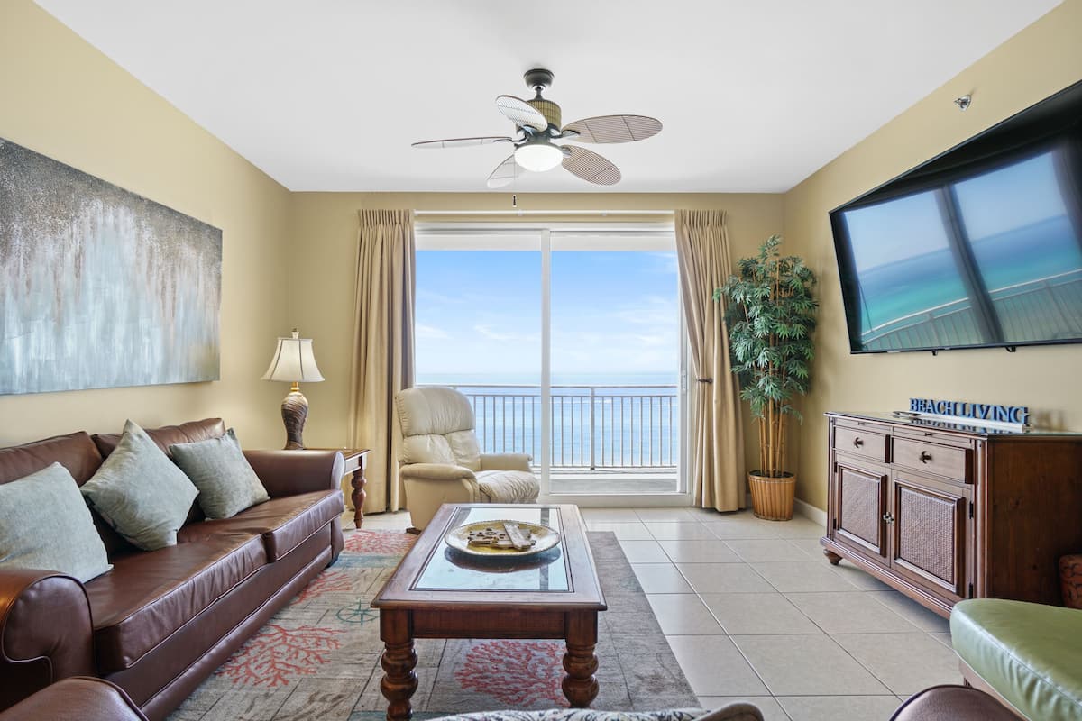 A living room with a brown leather sofa, a coffee table, a ceiling fan, a large window with a view of the ocean, and a television on the wall.