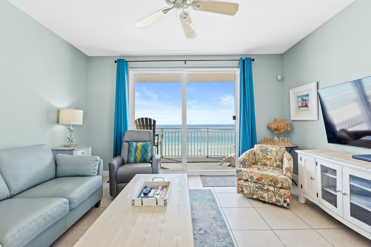 A living room with a sofa, armchair, coffee table, TV, and a balcony with a view of the beach.