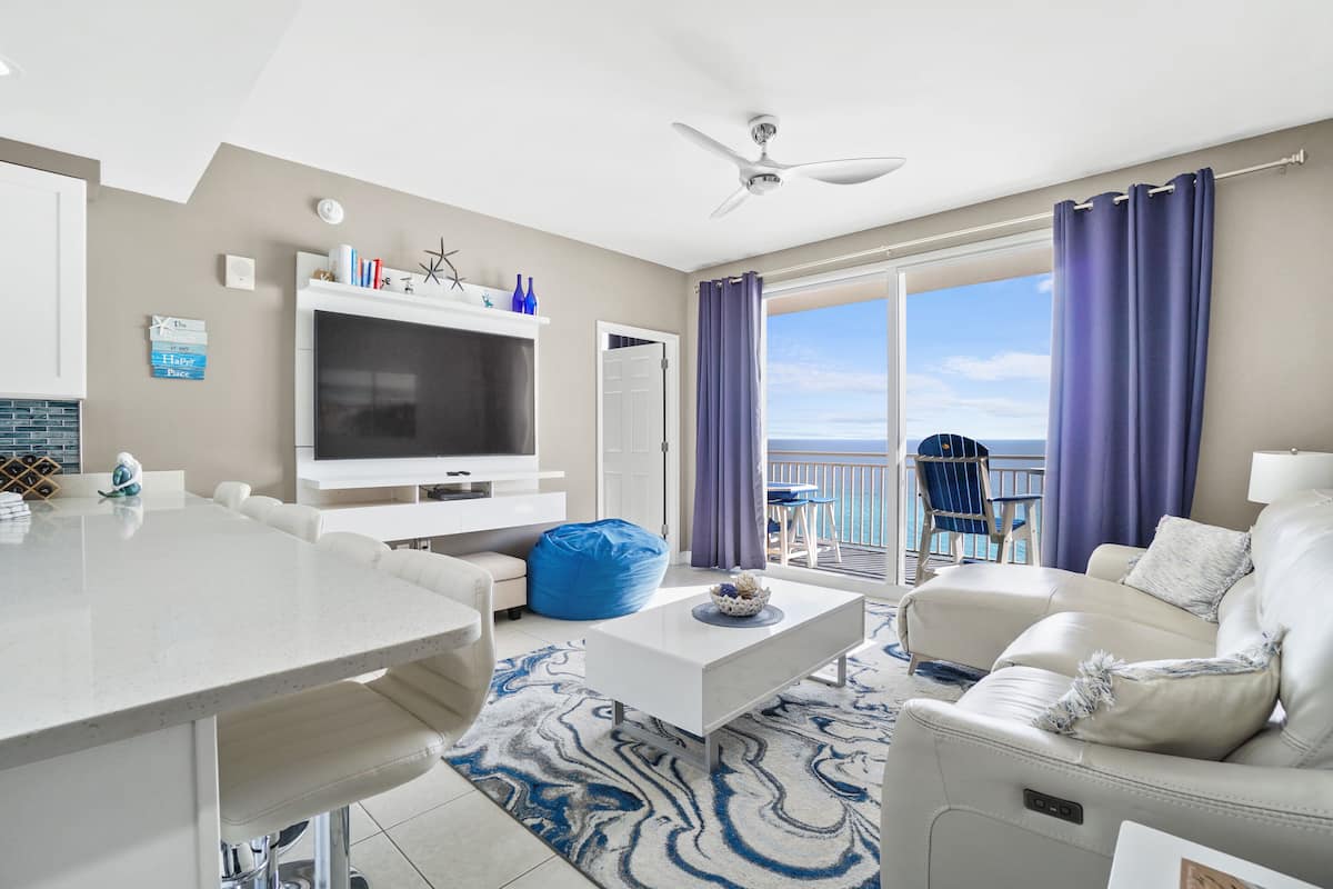A modern living room with a flat-screen TV, a white sofa, a coffee table, and a balcony with purple curtains.
