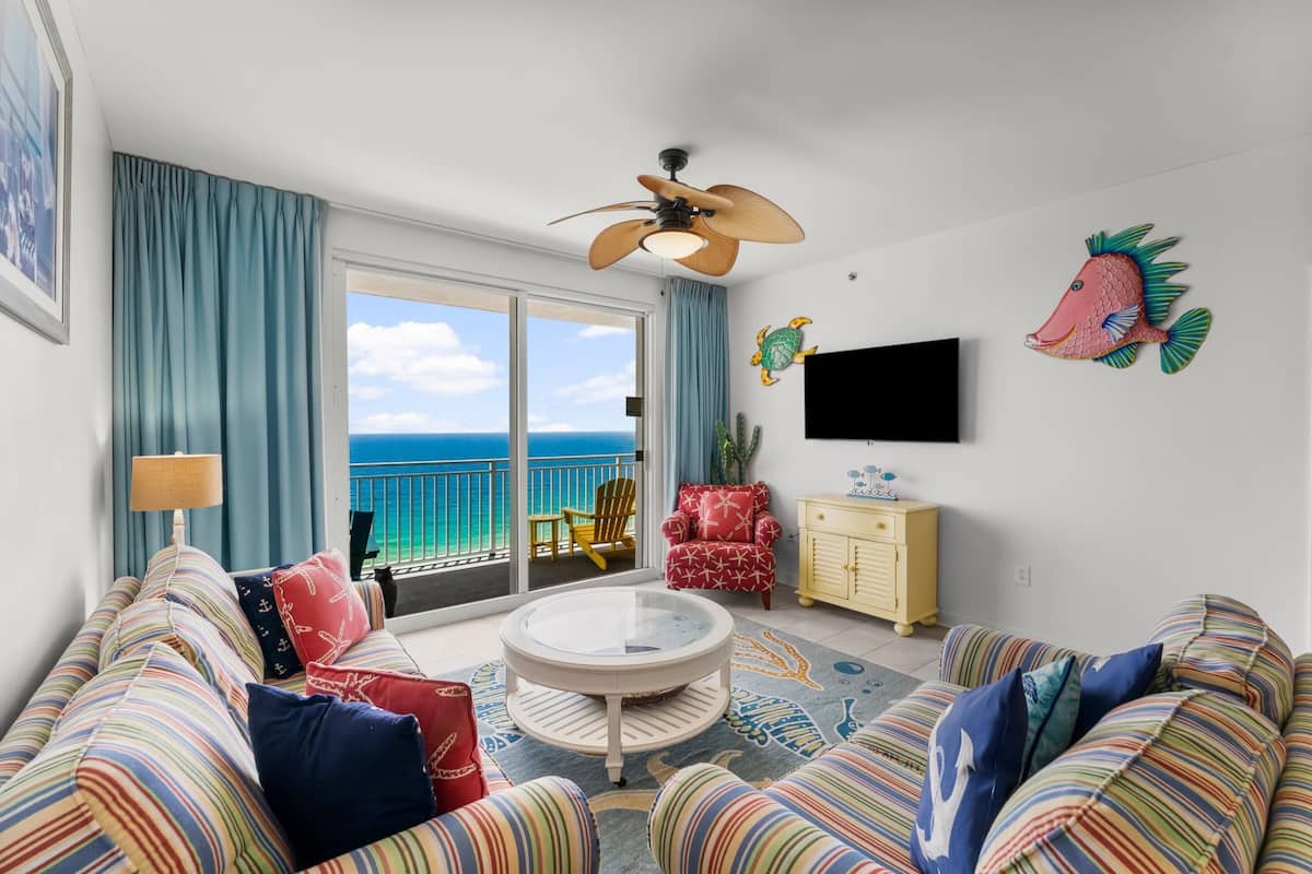 A living room with a view of the ocean, a ceiling fan, a flat-screen TV, and striped sofas.