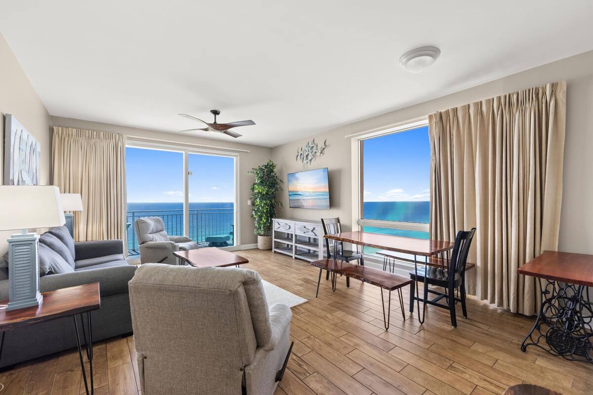 A spacious living room with a balcony, a ceiling fan, a TV, and a dining area with a view of the ocean.