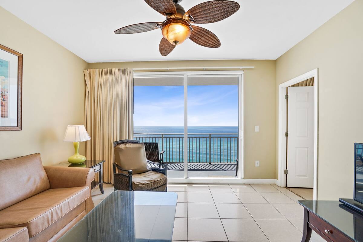 A hotel room with a balcony, a ceiling fan, a sofa, a table, and a TV.