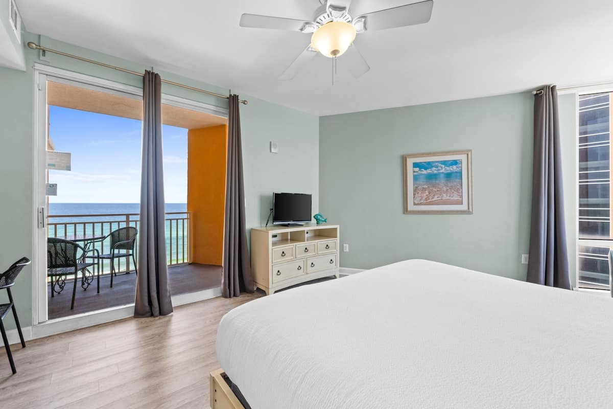 A bedroom with a bed, a dresser, a TV, and a balcony with chairs.
