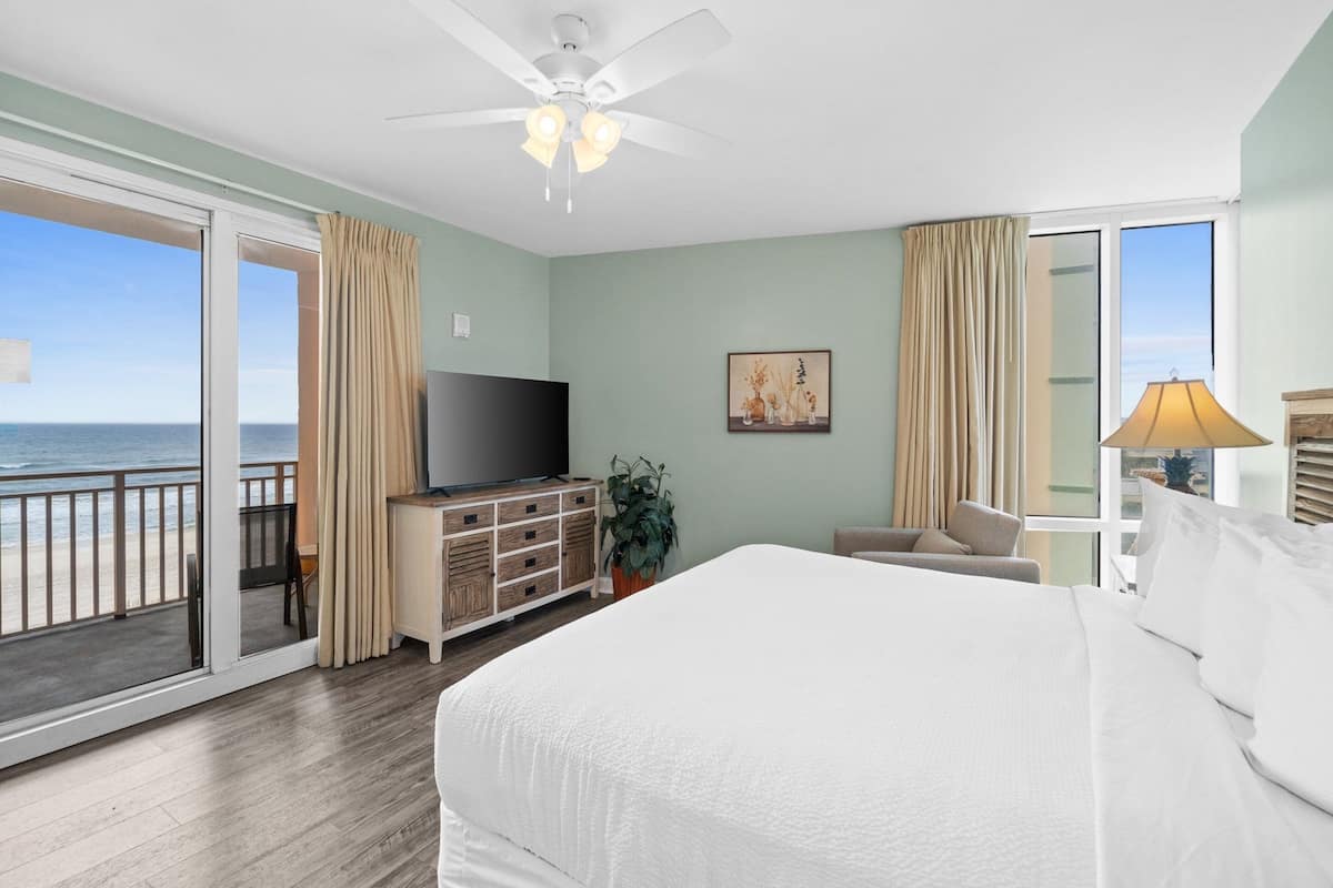 A hotel room with a large bed, a TV, a plant, and a balcony with a view of the ocean.