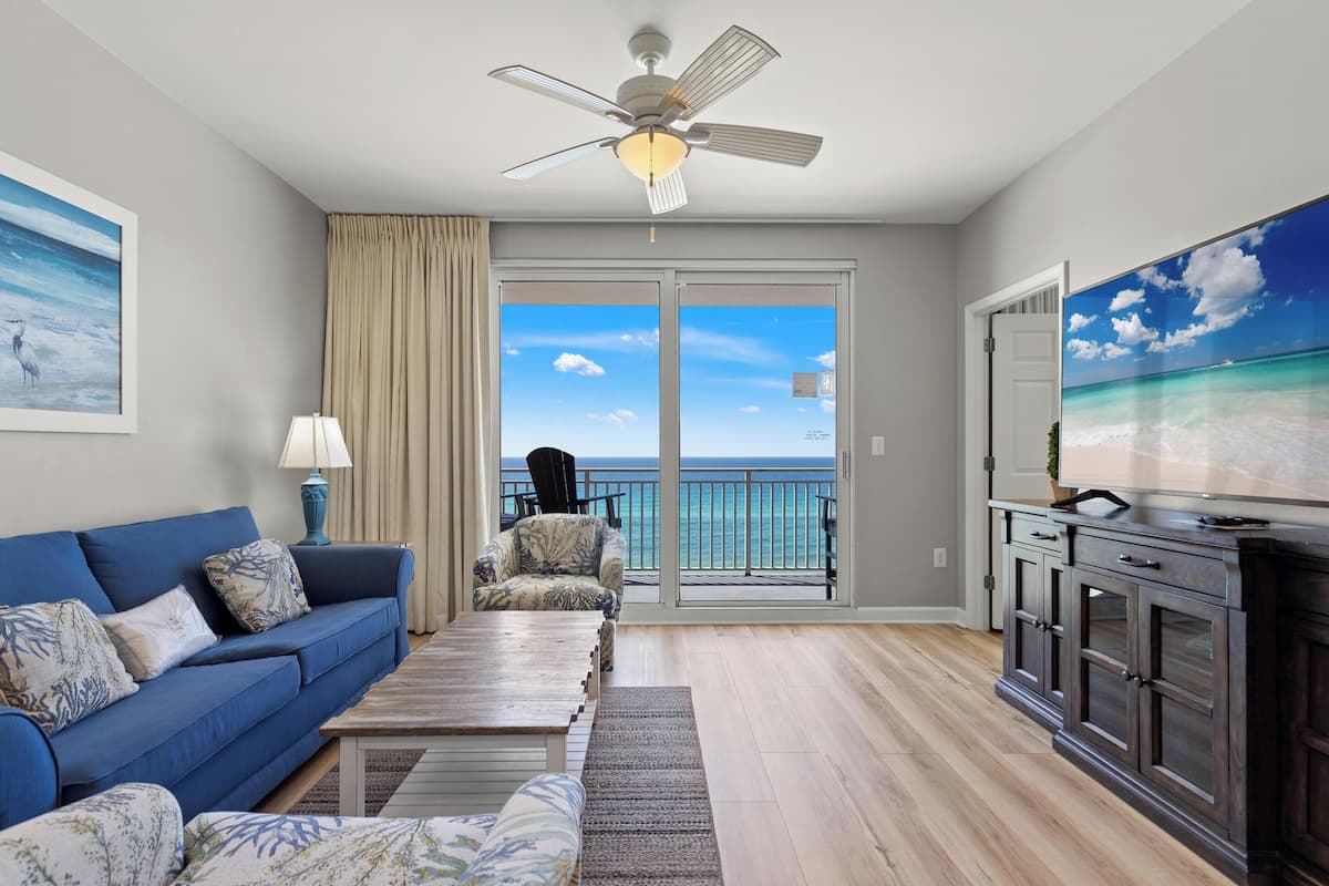 A living room with a blue sofa, a coffee table, a TV, and a view of the ocean through large glass doors.