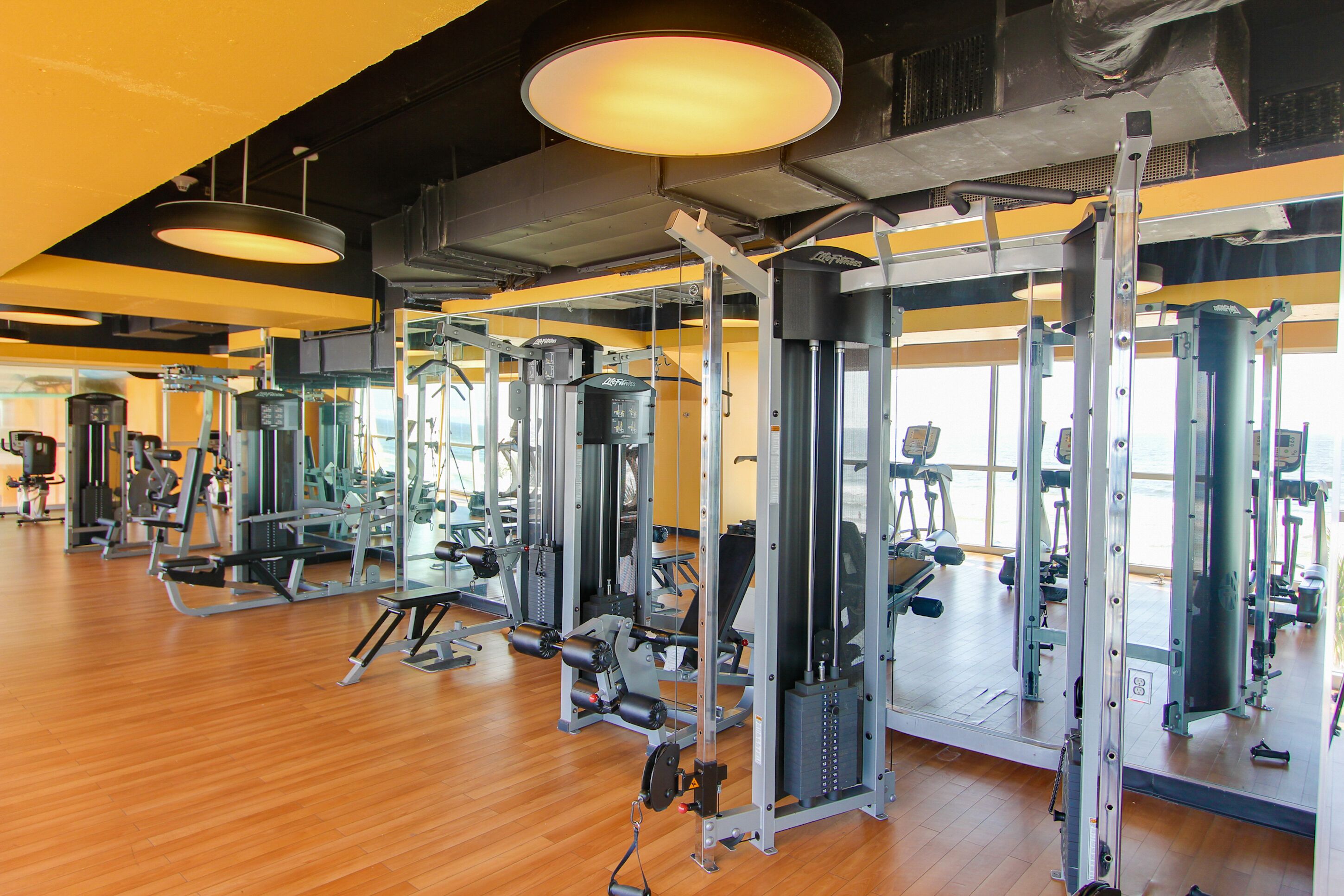 Ein Fitnessraum mit verschiedenen Trainingsgeräten, Holzboden und großen Fenstern für natürliches Licht.