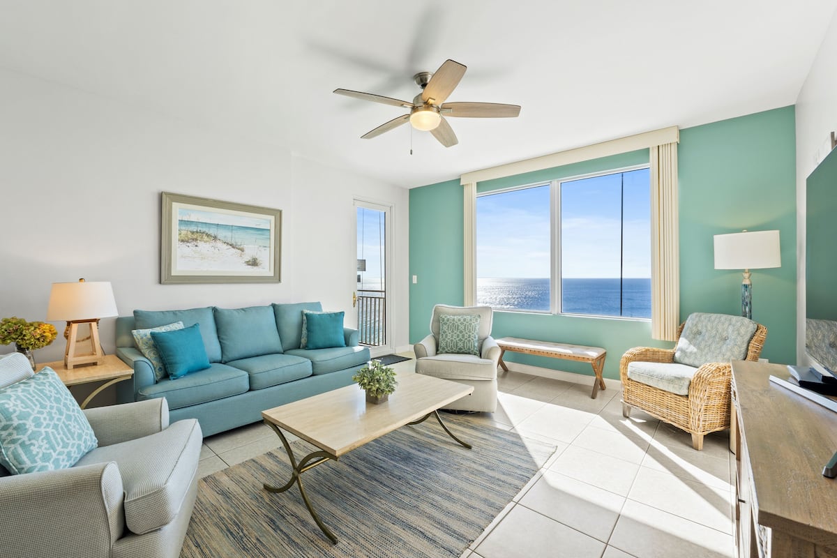 A spacious living room with a teal accent wall, a large window with a view of the ocean, a ceiling fan, and a variety of seating options including a sofa, chairs, and a bench.