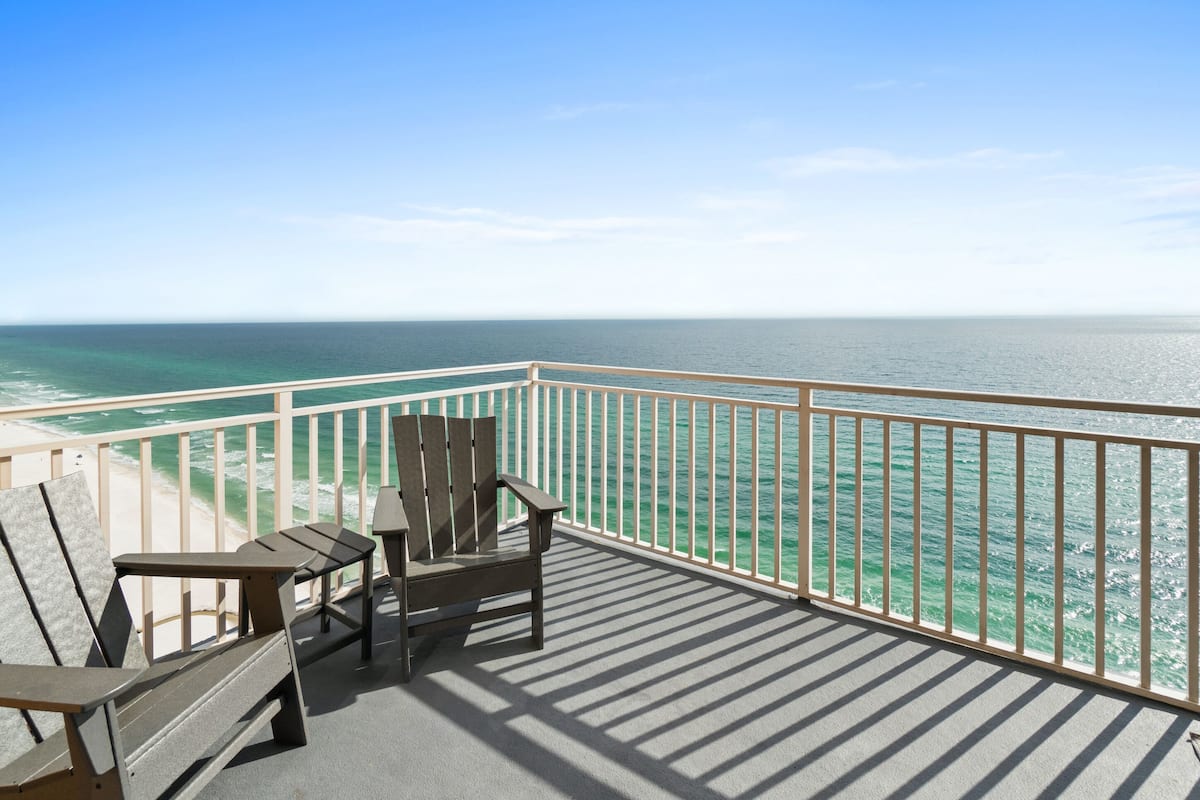 A balcony with two Adirondack chairs and a small table, overlooking a beach and the ocean.