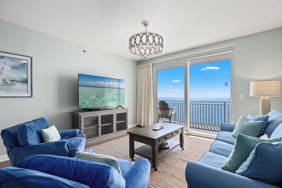 A living room with a large TV, a wooden coffee table, and a view of the ocean through large windows.