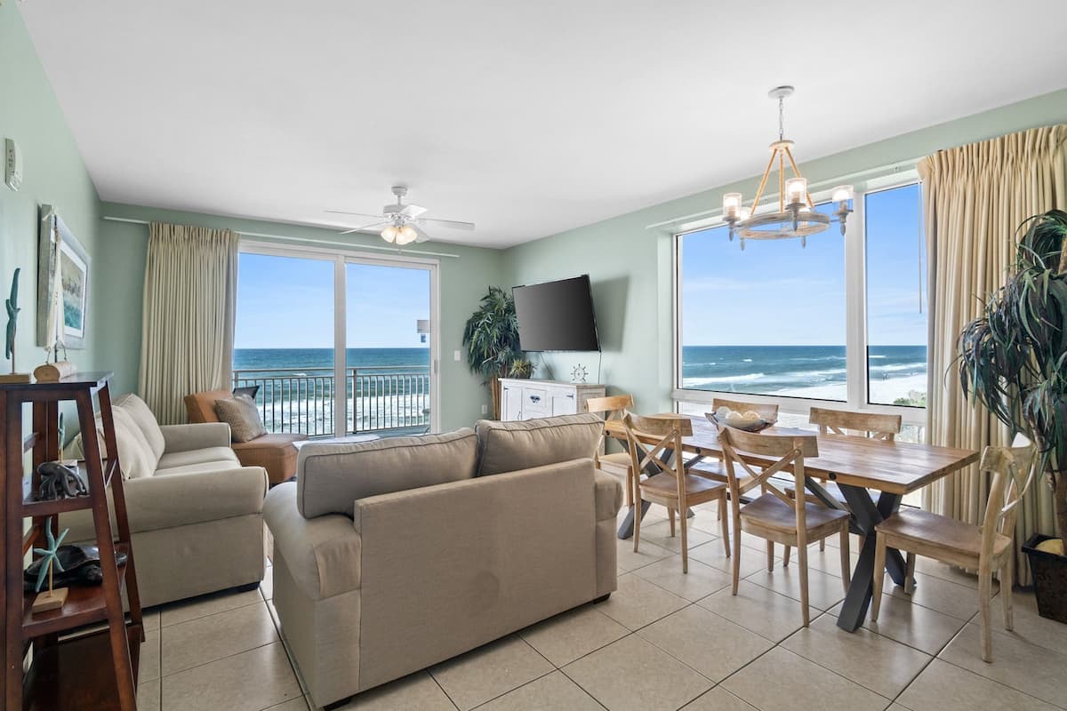A living room with a sofa, dining table, chairs, a TV, and a balcony with a view of the ocean.