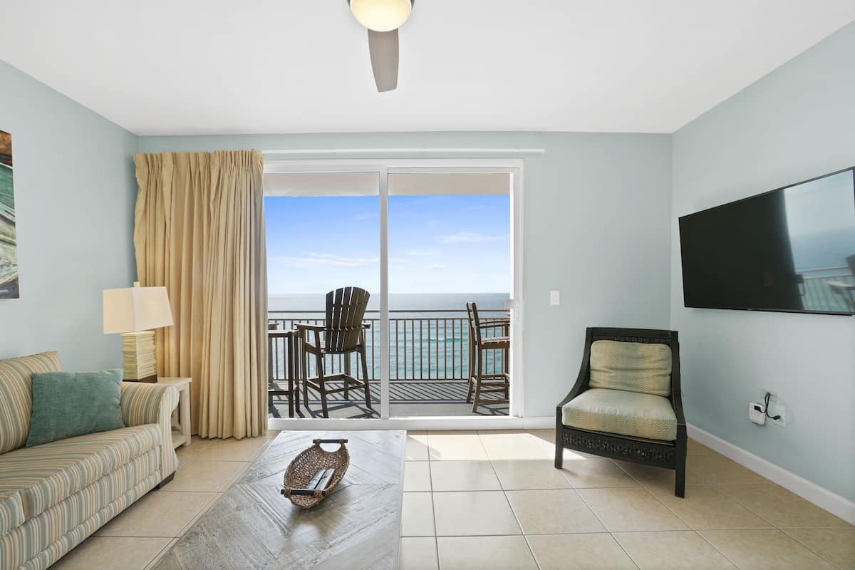 A room with a sofa, a coffee table, a chair, a TV, and a balcony with a view of the sea.