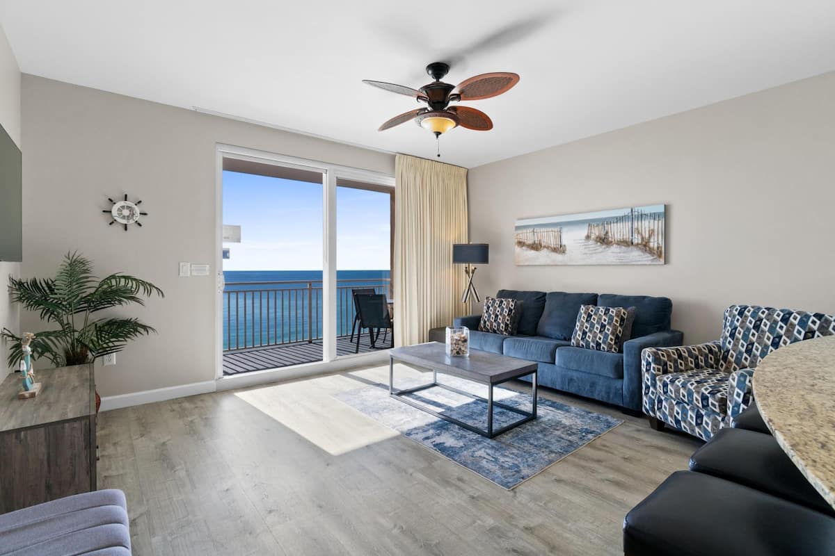 A spacious living room with a balcony, a ceiling fan, a sofa set, and a coffee table.
