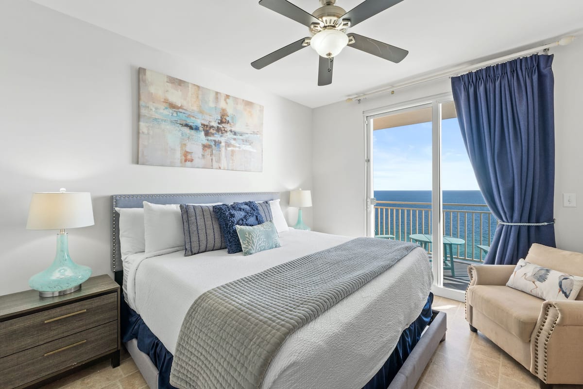 A bedroom with a bed, nightstands, a ceiling fan, a large window with curtains, and a view of the ocean.