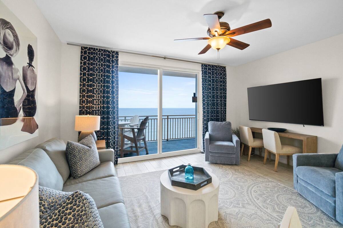 A modern living room with a sofa, coffee table, and TV, a balcony with a view of the sea, and a ceiling fan.