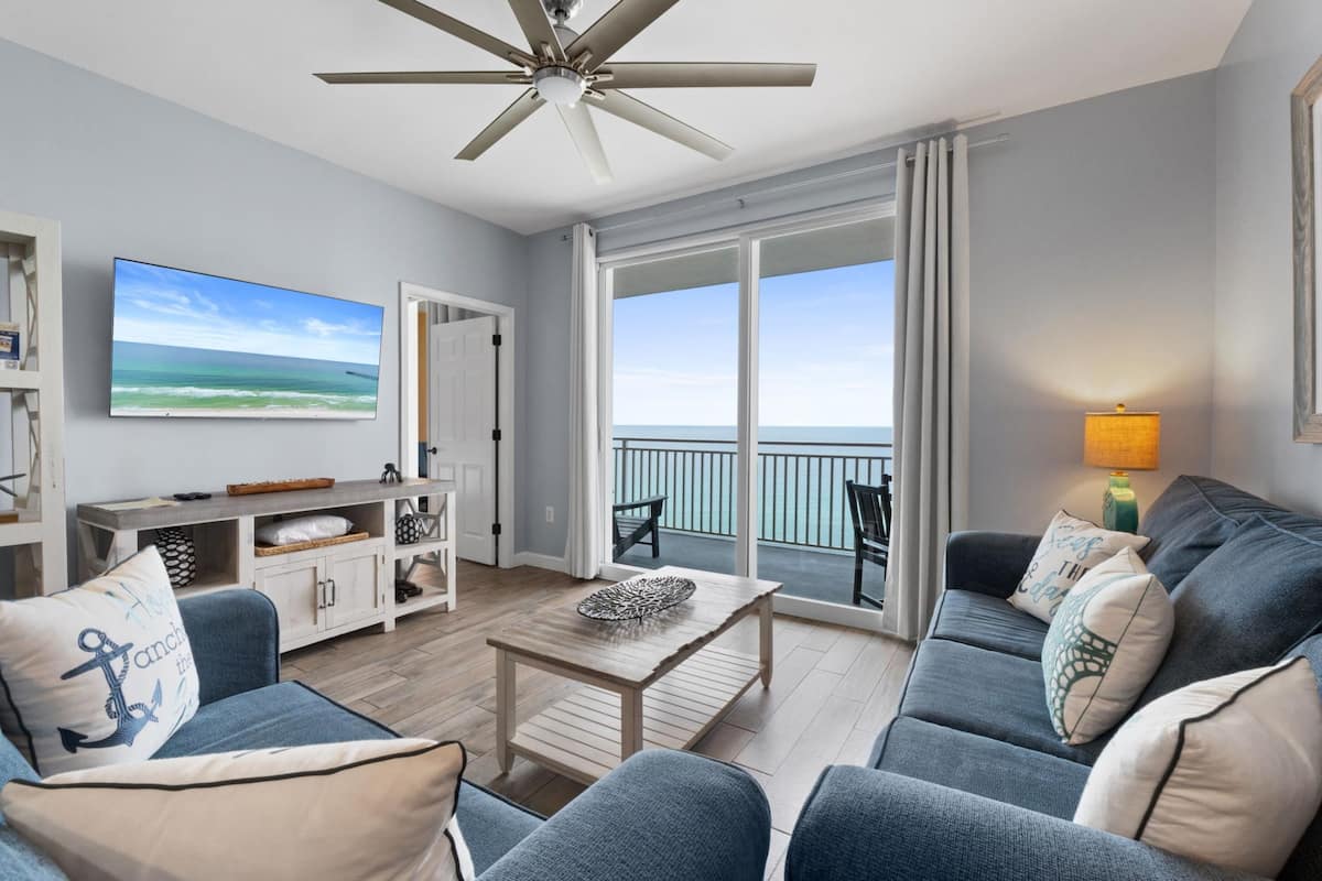 A spacious living room with a ceiling fan, a large TV, a balcony with a view, and a cozy seating area.