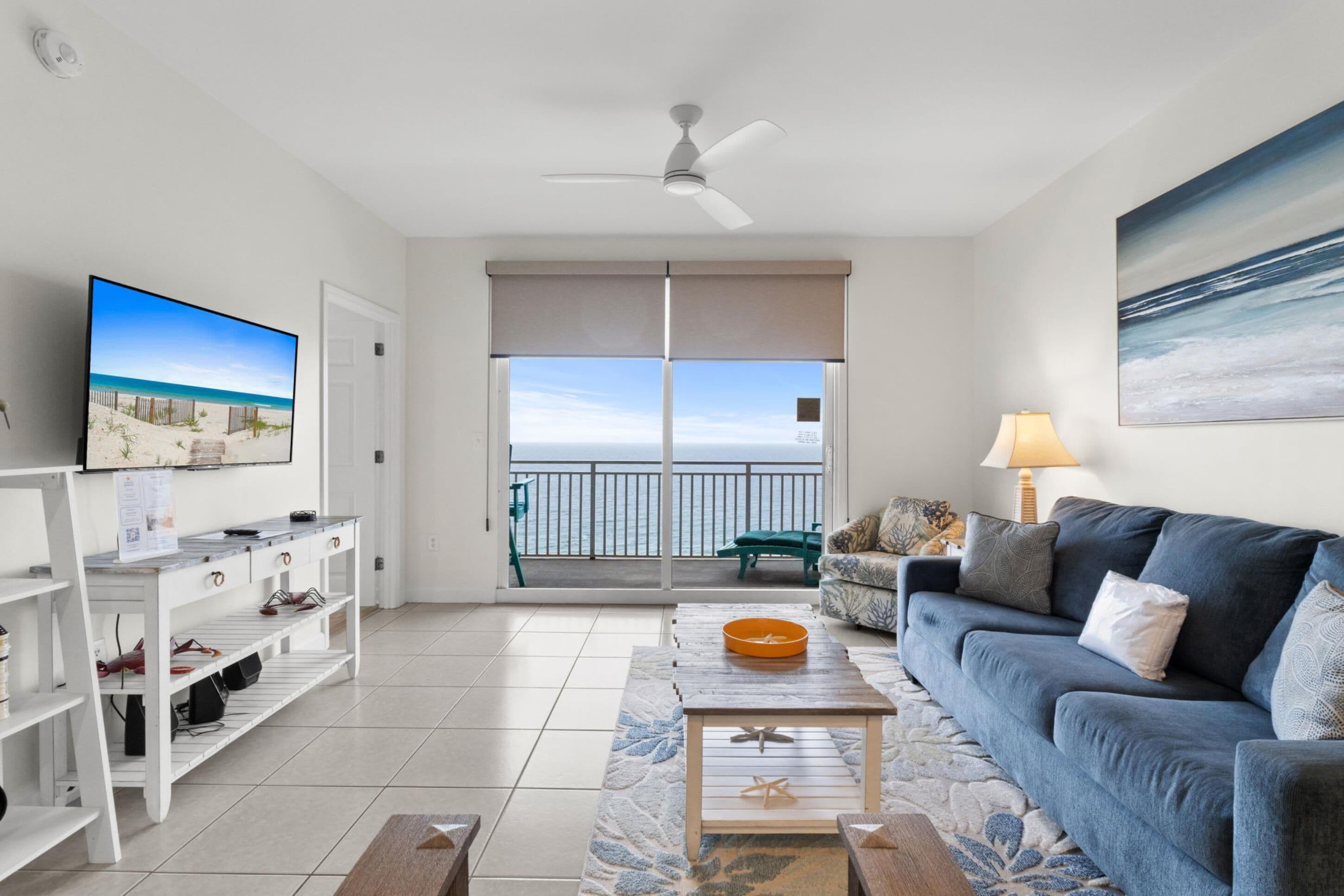 A living room with a TV, sofa, coffee table, and a balcony with a view of the ocean.
