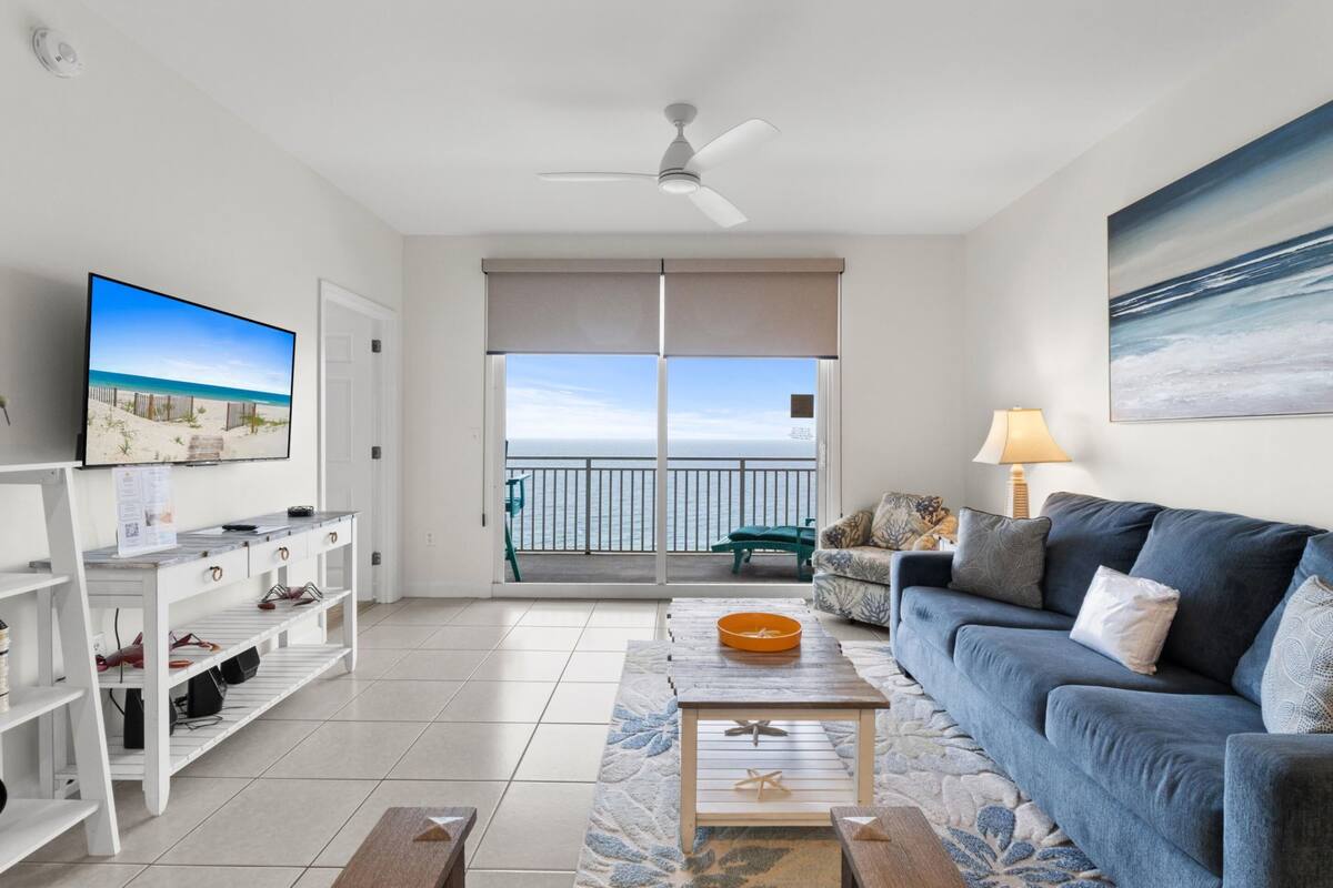 A living room with a TV, sofa, coffee table, and a balcony with a view of the ocean.