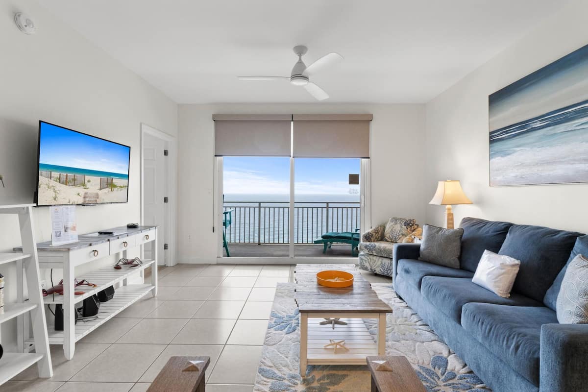 A living room with a TV, sofa, coffee table, and a balcony with a view of the ocean.