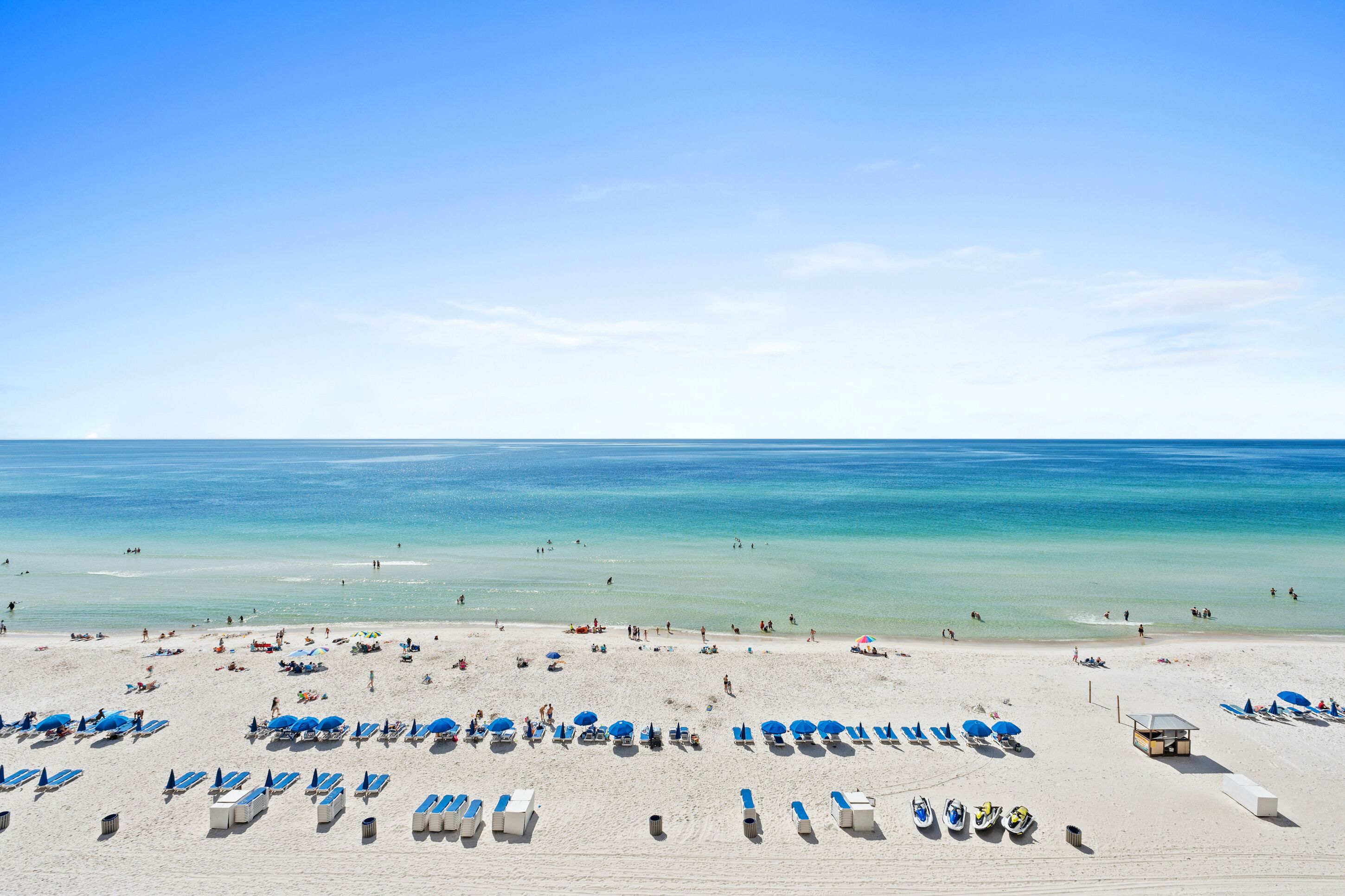 A beach with rows of sun loungers and umbrellas, a lifeguard tower, and clear blue water.
