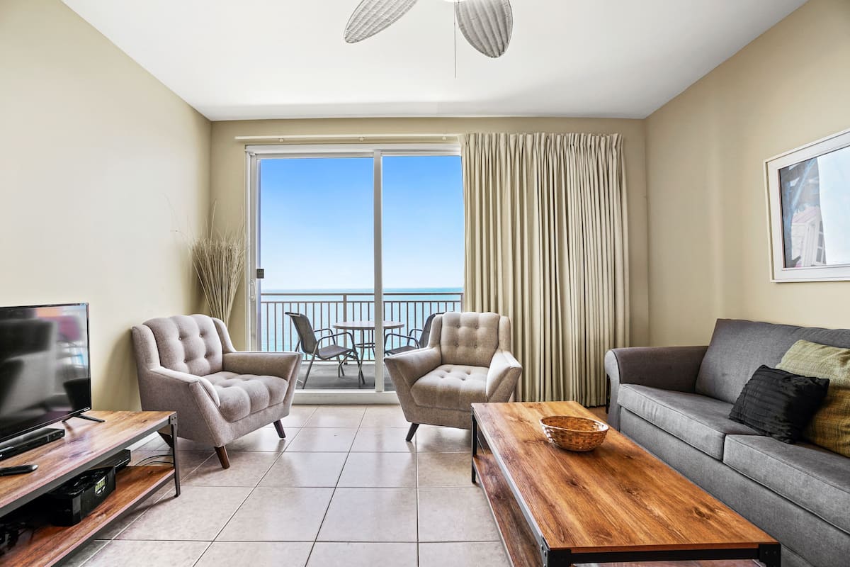 A living room with a TV, two armchairs, a sofa, a wooden coffee table, and a balcony with a view of the sea.