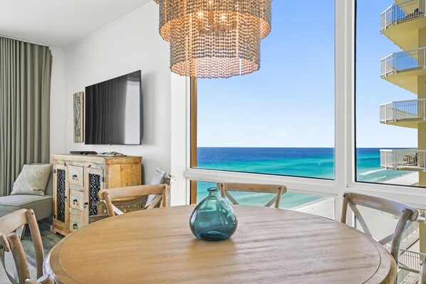 A room with a wooden dining table, a blue vase, a wicker chandelier, a flat-screen TV, and a view of the ocean.