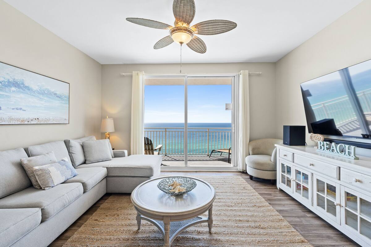 A modern living room with a sofa, a coffee table, a TV on a cabinet, and a balcony with a view of the ocean.