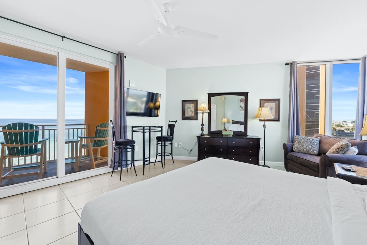 A hotel room with a large bed, a desk with two chairs, a TV, a dresser, and a balcony with chairs.