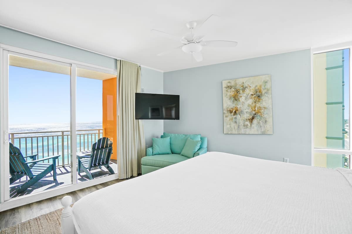 A bedroom with a large bed, a ceiling fan, a TV, a green couch, and a balcony with chairs and a view of the ocean.