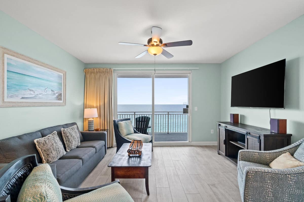 A living room with a sofa, armchairs, a coffee table, a TV, and a balcony with a view of the ocean.