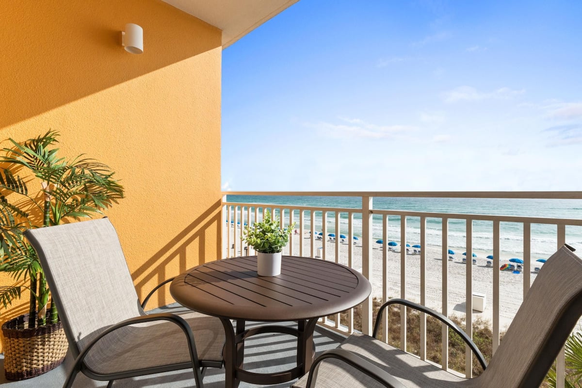 A balcony with a round table, two chairs, a potted plant, and a view of the beach.